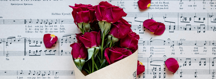 A bouquet of red roses sits on a sheet of music. Red rose petals are scattered across the sheet